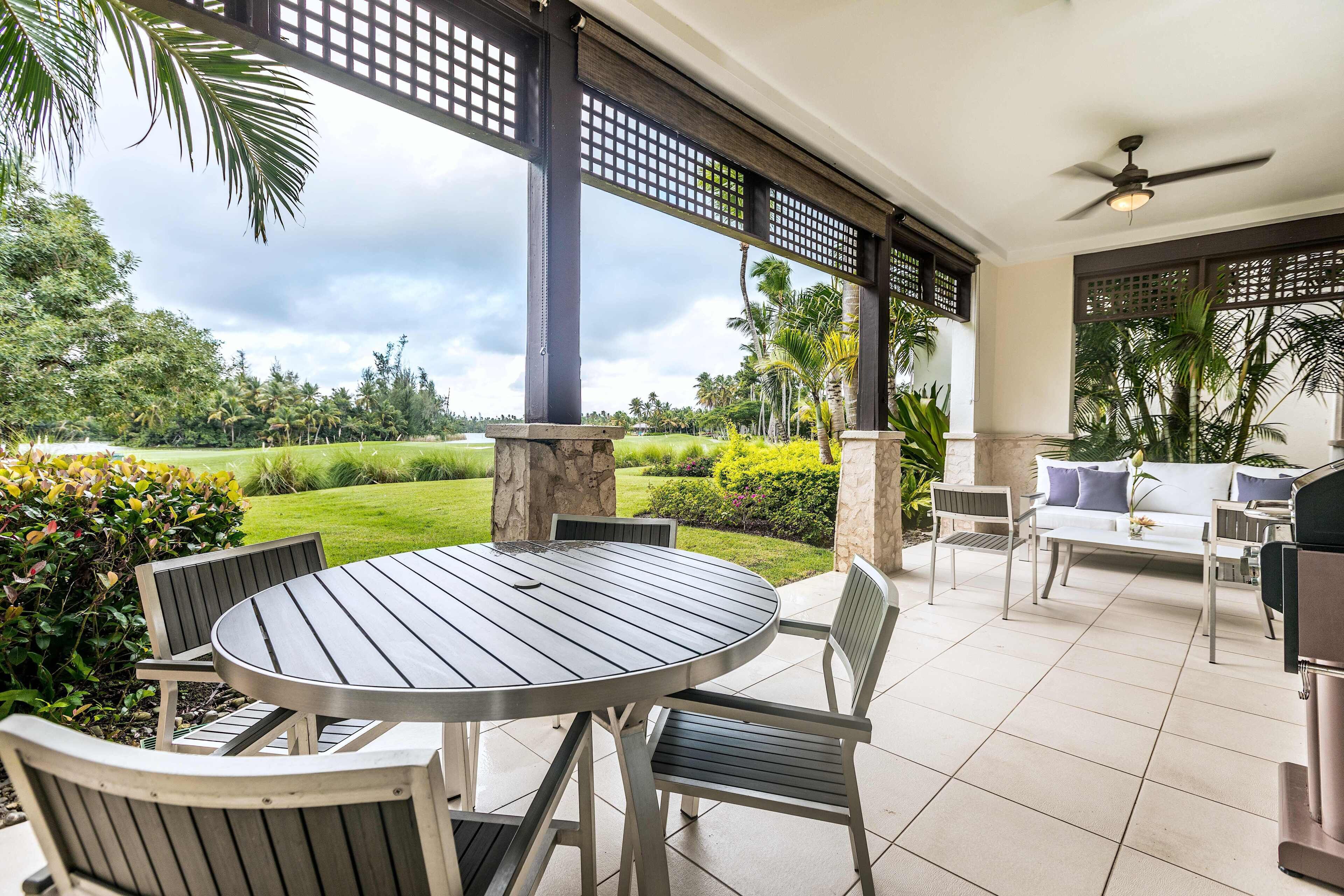 A covered patio area with a round table and chairs, a ceiling fan, and a view of a well-maintained garden.