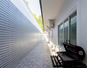 Ein schmaler Außenkorridor mit Grill und Bank, eine weiße Wand mit horizontalen Rollläden und einem Fenster mit Jalousien.