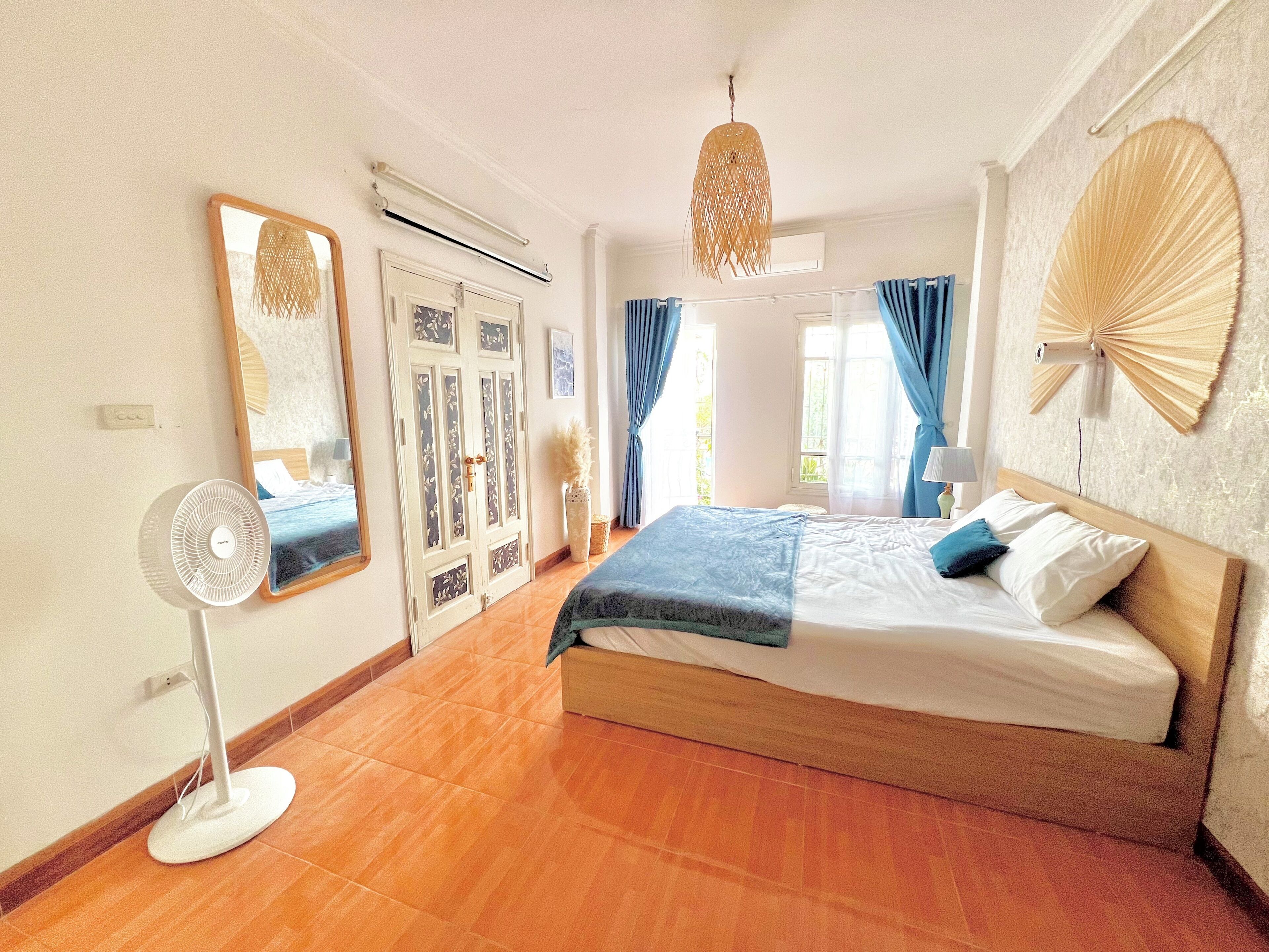 A bedroom with a bed, a standing fan, a large mirror, a door with decorative panels, and a wall fan.