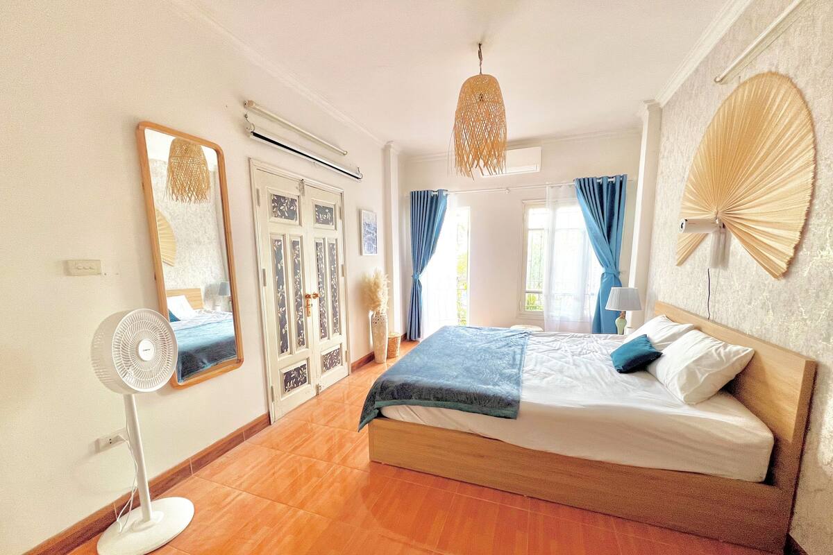 A bedroom with a bed, a standing fan, a large mirror, a door with decorative panels, and a wall fan.