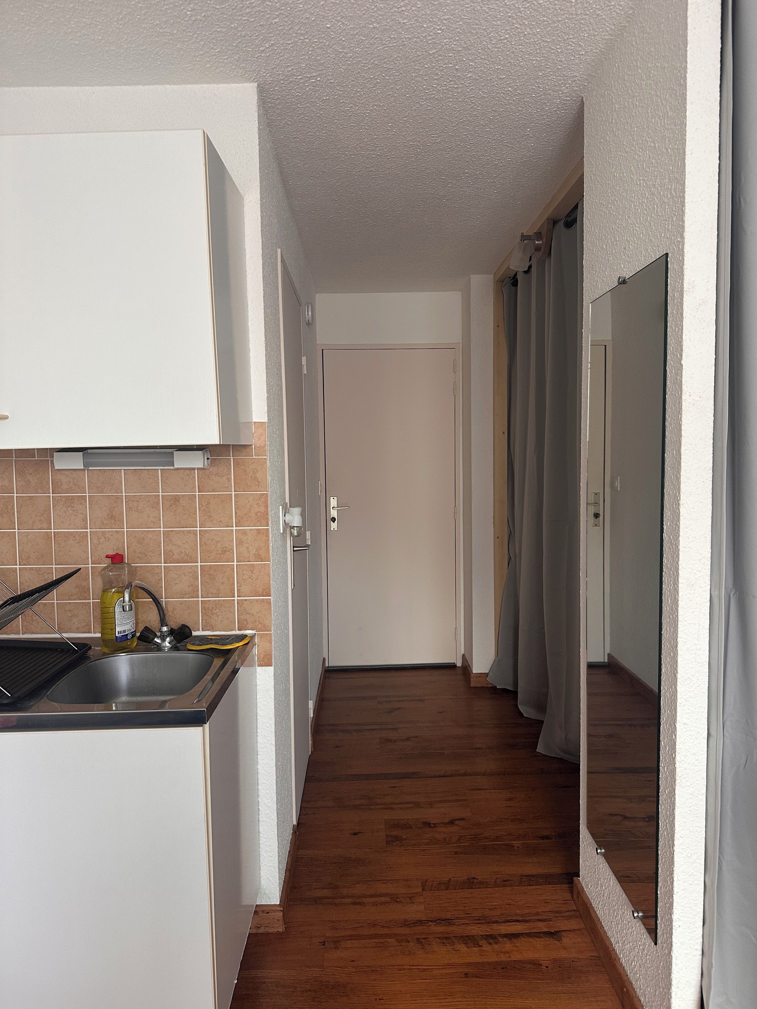 Un couloir étroit, avec une cuisine d’un côté, un plancher en bois et une porte au fond.