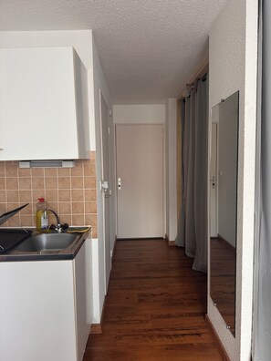 Un couloir étroit, avec une cuisine d’un côté, un plancher en bois et une porte au fond.