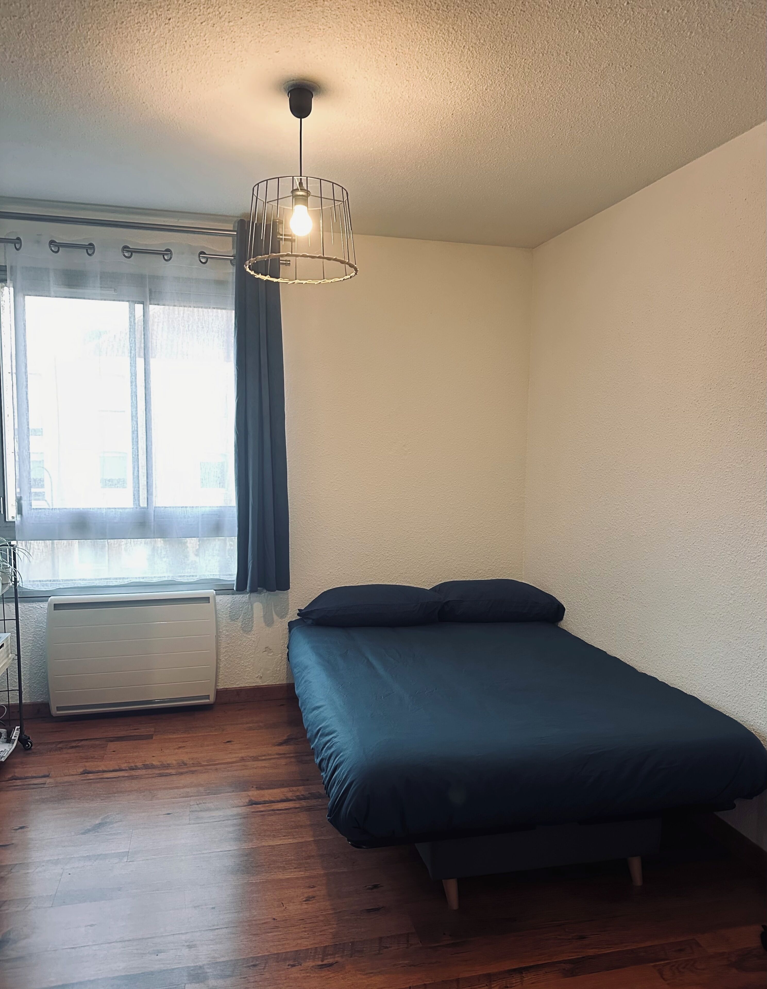 Une pièce comprenant un lit, un radiateur, une fenêtre avec des rideaux et un luminaire suspendu.