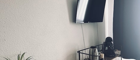 Een kamer met een flatscreen televisie aan de muur, een houten eettafel met een potplant, een koffieapparaat, en een klein karretje met spullen.