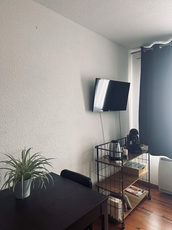 Une pièce dotée d’un téléviseur à écran plat fixé au mur, d’une table à manger en bois agrémentée d’une plante en pot, d’une machine à café et d’un petit chariot contenant divers objets.