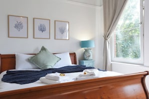 Ein Bett mit Holz-Kopfstütze, einem grünen Kopfkissen, einer blauen Decke und einem weißen Handtuch. An der Wand hängen drei gerahmte Bilder, auf einem Nachttisch steht eine Lampe und ein Fenster mit Vorhängen.