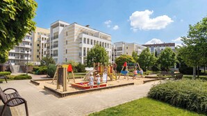 Ein Spielplatz mit Rutschen und Klettergerüsten, umgeben von einem befestigten Bereich mit Bänken und Grünflächen.