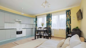 Ein Schlafzimmer mit Bett, einem Esstisch mit Stühlen, einer Küchenzeile inklusive Schränken und Geräten sowie einem Fenster mit Vorhängen.