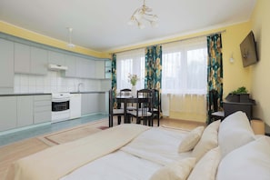 Ein Schlafzimmer mit Bett, einem Esstisch mit Stühlen, einer Küchenzeile inklusive Schränken und Geräten sowie einem Fenster mit Vorhängen.