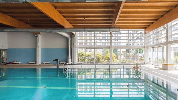 Ein Hallenbad mit klarem, blauem Wasser, Holzbalken an der Decke und großen Fenstern mit Blick auf ein Wohngebäude.
