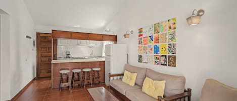 Ein Wohnzimmer mit einer Couch und Holzmöbeln, eine Küche mit weißen Schränken und einer Kaffeemaschine, sowie eine Wand mit farbenfroher Kunst.