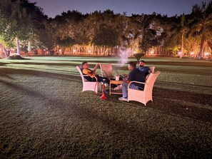 Area esterna con posti a sedere serale, con sedie in vimini e un tavolo, circondata da alberi e illuminata da luci soffuse.