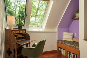 A room with a sloped ceiling, a desk with drawers, a chair, a bookshelf, and a window with a view of trees.