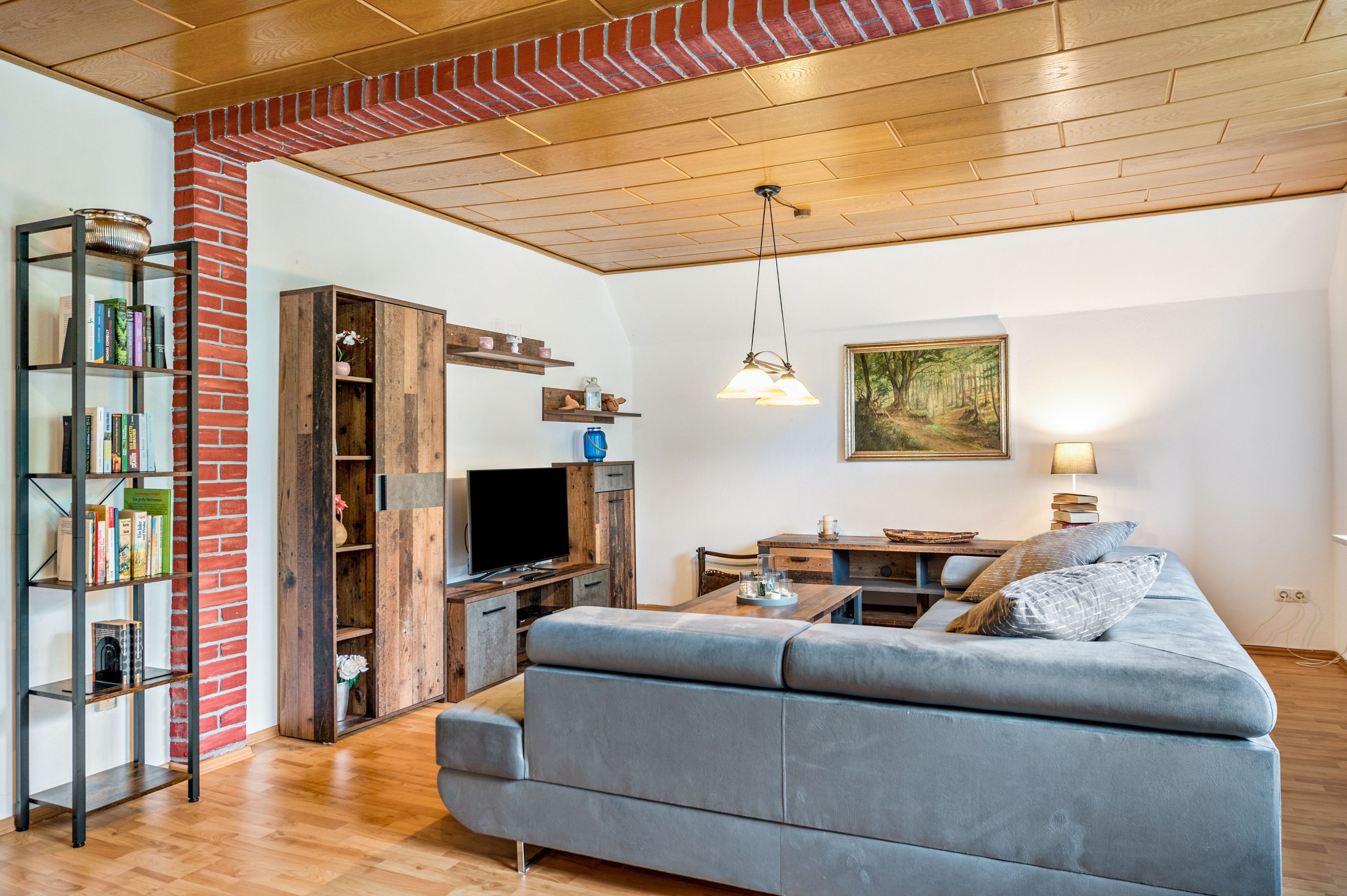 A modern living room with a grey sofa, wooden flooring, a TV, and a bookshelf.
