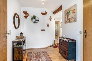 Un couloir doté d’un plancher en bois, d’un miroir rond, d’une étagère murale ornée d’une plante, d’un tapis à motifs et d’une commode en bois avec des tiroirs.