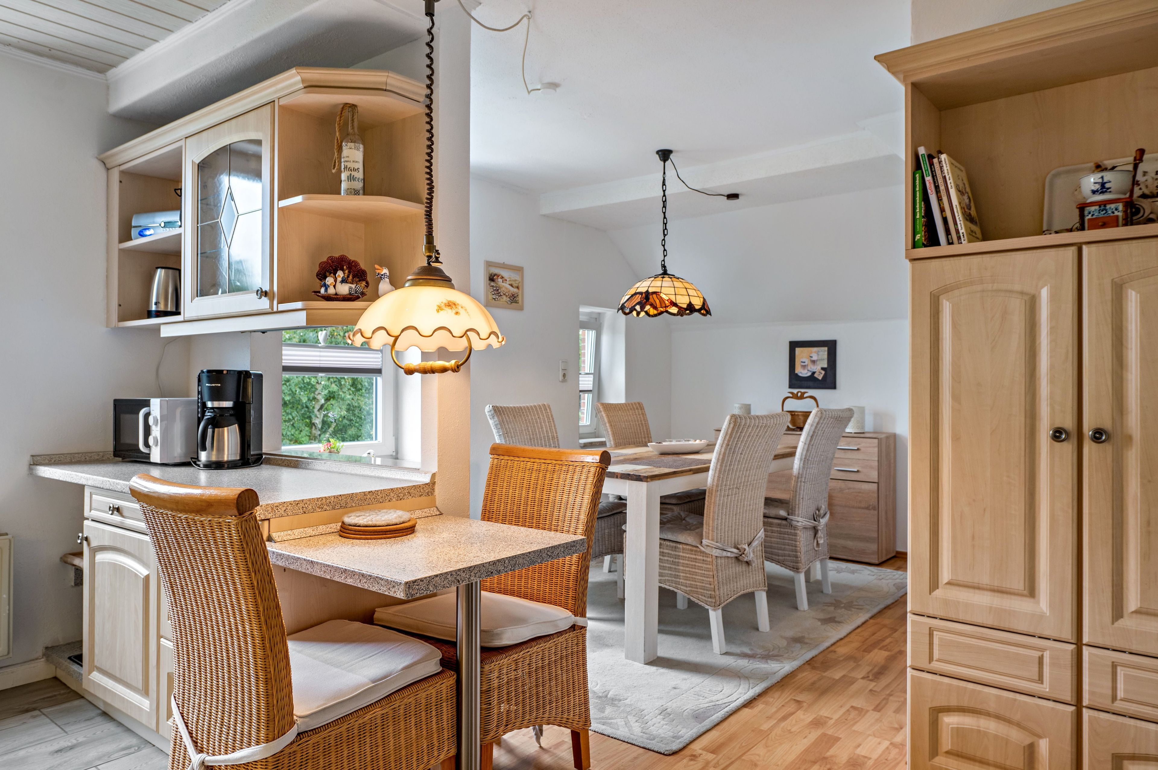 A modern kitchen with a dining area, featuring wicker furniture, a coffee machine, and pendant lighting.