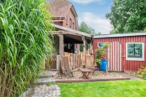 Ein rustikales Haus mit einer Holzdielenveranda, einem roten Geräteschuppen und einer großen, grünen Pflanze.