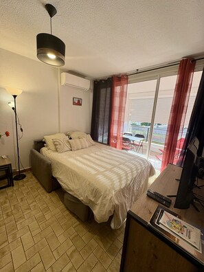 A bedroom with a bed, a desk with a computer, a floor lamp, and a window with curtains. - Well-located, quiet studio with a pool (Gruissan)