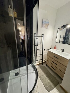 A modern bathroom with a glass shower enclosure, a wooden vanity with a white sink, and a mirror. - Well-located, quiet studio with a pool (Gruissan)