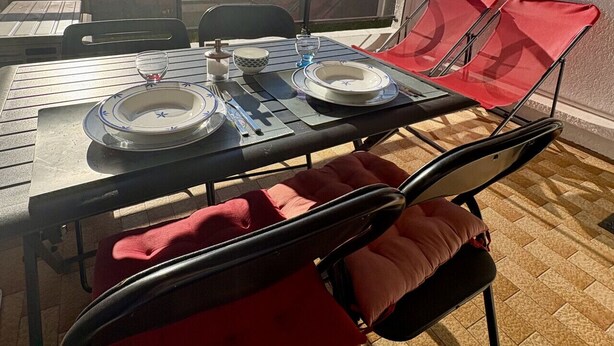 A dining area with a table set for two, red chairs, and a view of buildings through a large window. - Well-located, quiet studio with a pool (Gruissan)