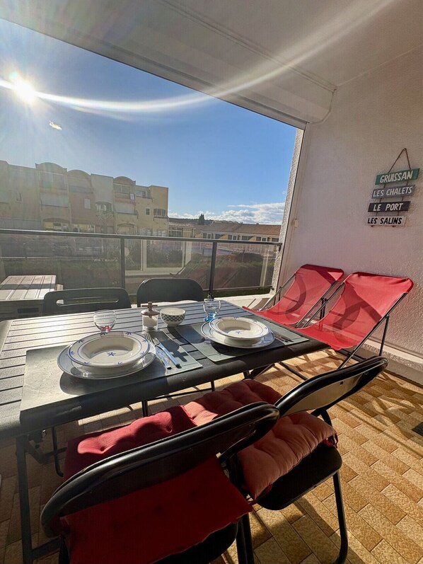 A dining area with a table set for two, red chairs, and a view of buildings through a large window. - Well-located, quiet studio with a pool (Gruissan)