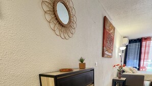 A living room with a wooden cabinet, a decorative mirror, a small plant, a sofa, and a window with curtains. - Well-located, quiet studio with a pool (Gruissan)