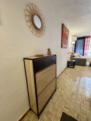 A living room with a wooden cabinet, a decorative mirror, a small plant, a sofa, and a window with curtains. - Well-located, quiet studio with a pool (Gruissan)