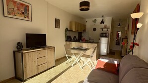 A living room with a sofa, a TV, a wooden cabinet, and a kitchen area in the background. - Well-located, quiet studio with a pool (Gruissan)