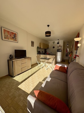 A living room with a sofa, a TV, a wooden cabinet, and a kitchen area in the background. - Well-located, quiet studio with a pool (Gruissan)