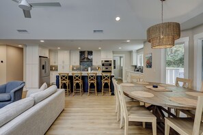 A modern kitchen with white cabinets, a central island with bar stools, a dining table with placemats, and a ceiling fan. - 3 Jessamine - A Coastal Comfort Escape Available Starting August 2026! (Hilton Head Island)