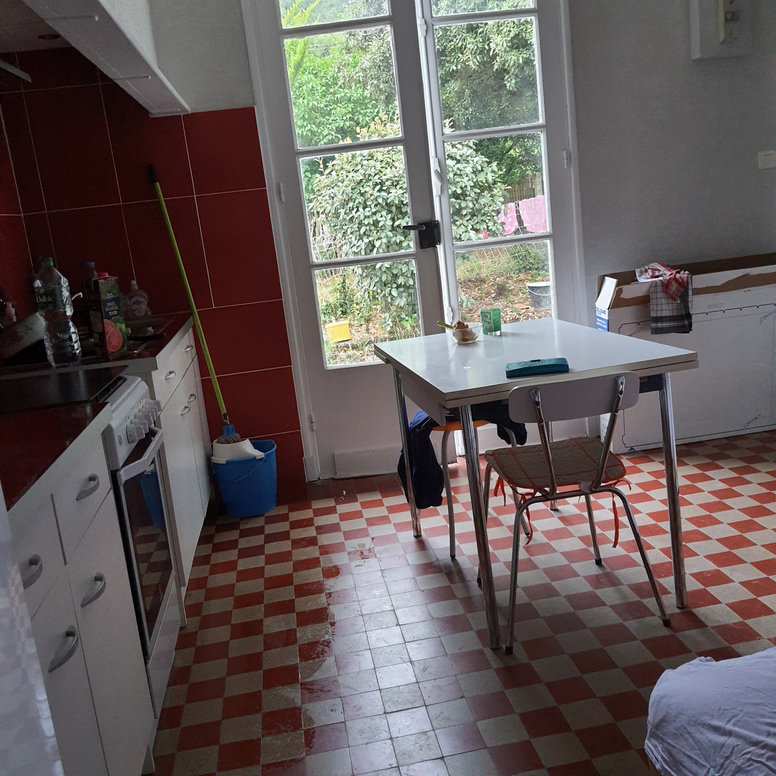 A kitchen with a checkered floor, a window with a view of greenery, a table with a chair, and a mop leaning against the wall.