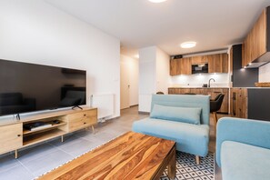 A modern living room with a flat-screen TV, wooden coffee table, blue sofa, and a kitchen area with wooden cabinets and a microwave. - CIMES B105C  By Avecoeur (Briançon)
