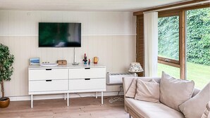 En stue med en hvid sofa, et fjernsyn monteret på væggen, et lille bord med en lampe og et vindue med udsigt til grønne områder.