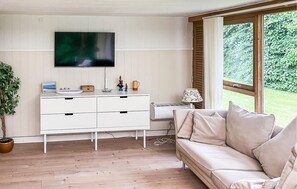 A living room with a white sofa, a TV mounted on the wall, a small table with a lamp, and a window with a view of greenery. - 4 bedroom gorgeous home in Højslev (Højslev)