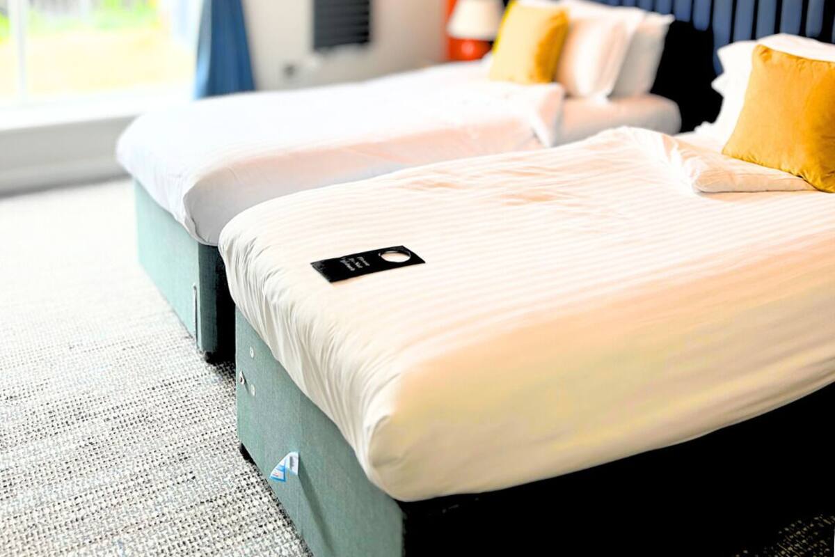 Habitación de hotel con dos camas, un ventanal con cortinas, un ventilador de techo y un cuadro en la pared.