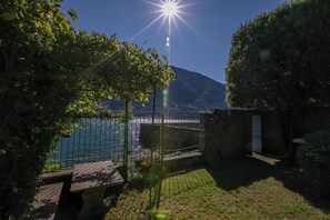 Ein Innenhof mit einer Steinsitzbank, einem Metallzaun und Blick auf ein Gewässer und Berge.