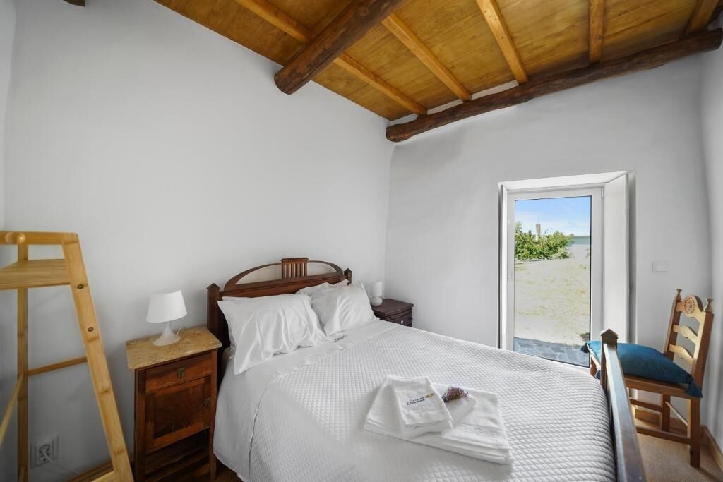 A bedroom with a wooden bed, a nightstand, a bedside table with a lamp, a wooden ladder, and a view of the outdoors through a door.