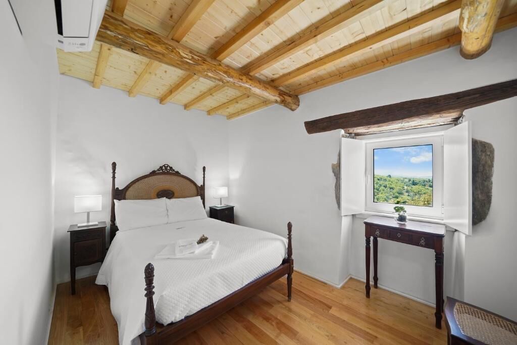 Une chambre avec un plafond en bois, un lit doté d’une tête de lit en bois, une table de chevet, une commode, une fenêtre donnant sur un paysage verdoyant et une petite table avec une plante.