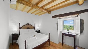 Une chambre avec un plafond en bois, un lit doté d’une tête de lit en bois, une table de chevet, une commode, une fenêtre donnant sur un paysage verdoyant et une petite table avec une plante.