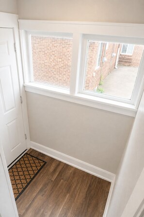 Un couloir avec un plancher en bois, une porte blanche et une fenêtre donnant sur un bâtiment en briques.