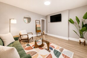 Ein Wohnzimmer mit Sofa, Sessel, Couchtisch, Stehlampe, Spiegel, Fernseher und einer Zimmerpflanze.
