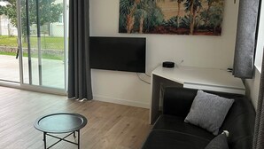 A modern living room with a sofa, a small round table, a flat-screen TV mounted on the wall, and a painting of a tropical landscape.