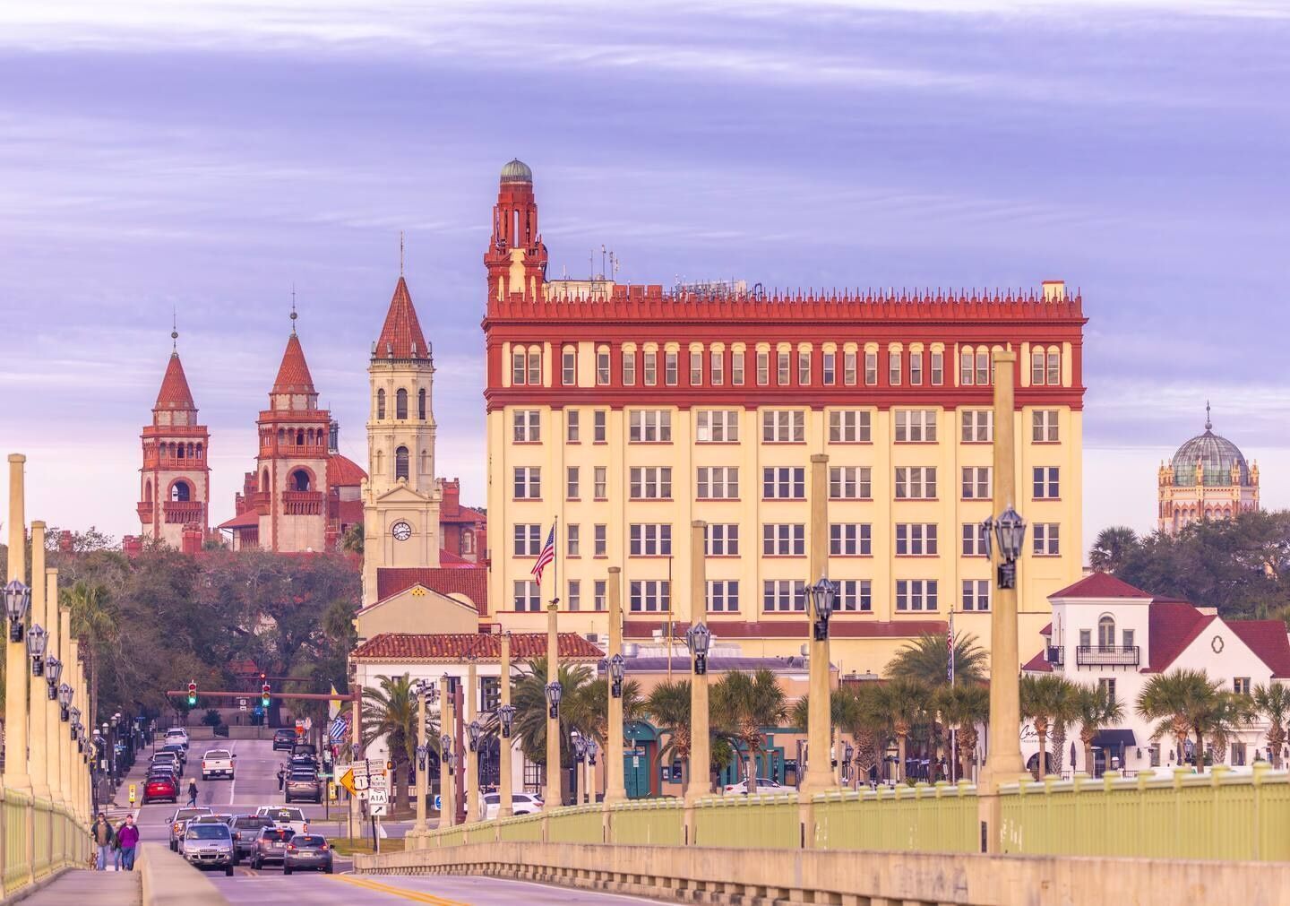 A large building with a red roof and multiple towers, a yellow building with a dome, and a bridge with cars and pedestrians.