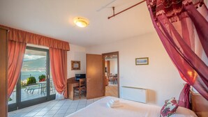 A hotel room with a bed, a desk, a chair, a balcony with a view, and a door leading to another room. - ApartmentsGarda - Villa Giada - RUBINO (TORRI DEL BENACO)