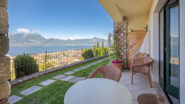 A balcony with a view of a lake and mountains, wicker chairs, a table, and a stone wall. - ApartmentsGarda - Villa Giada - RUBINO (TORRI DEL BENACO)
