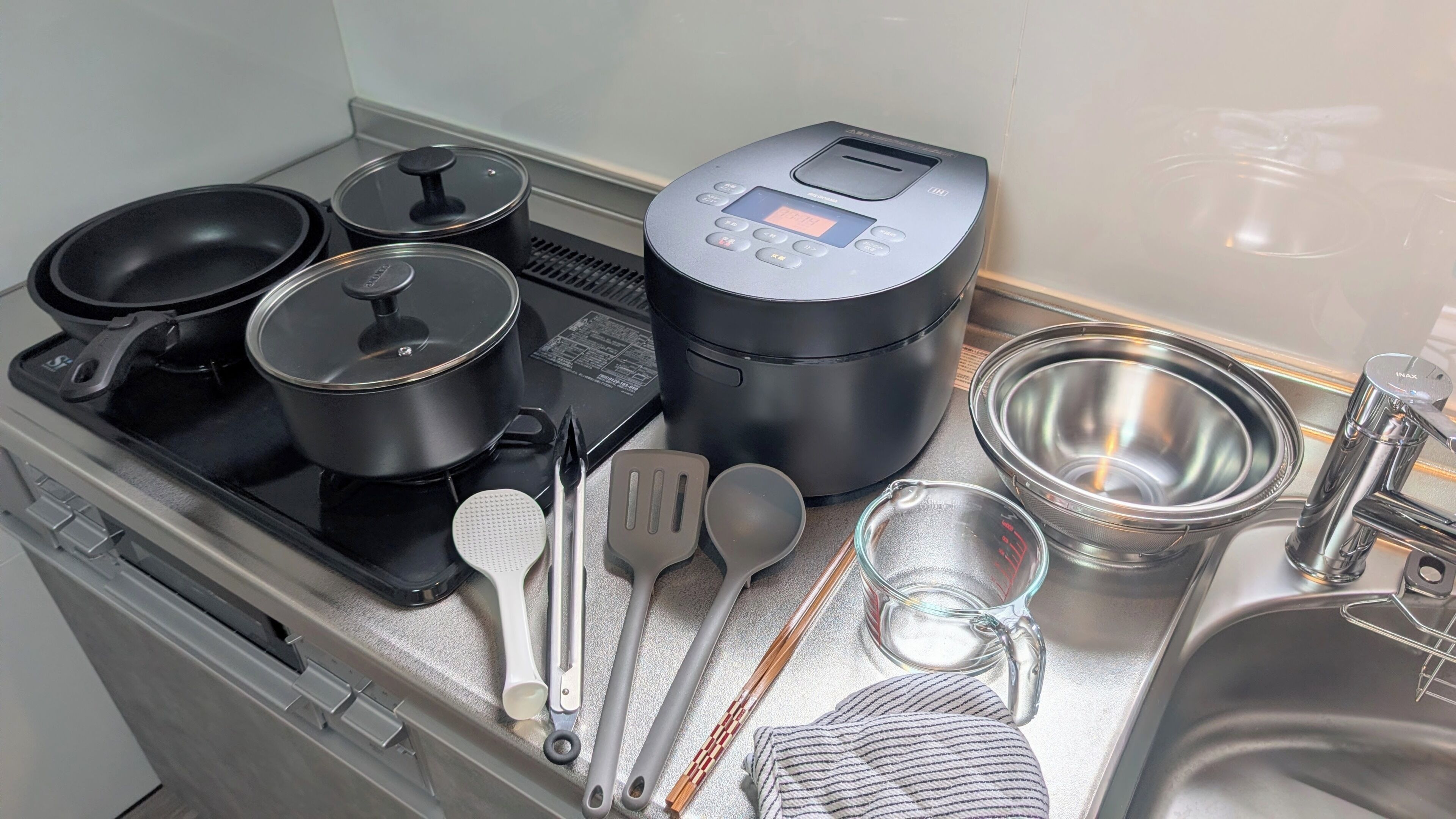 A compact kitchen with a stove, pots, a rice cooker, and various utensils.