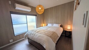 Un dormitorio con una cama grande, una ventana con vidrio esmerilado, aire acondicionado, una lámpara colgante y una mesita de noche con lámpara.