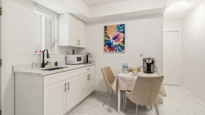 Een kleine keuken met witte kasten, een magnetron, een koffiezetapparaat en een eettafel met stoelen.
