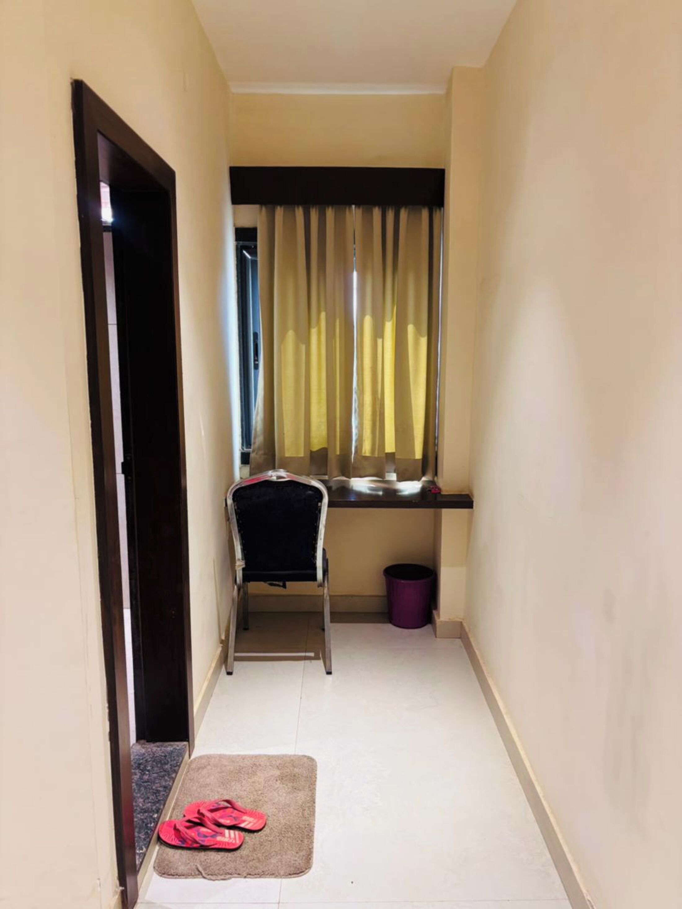 A narrow hallway with a chair, a small window with curtains, a trash bin, and a pair of slippers on a mat.