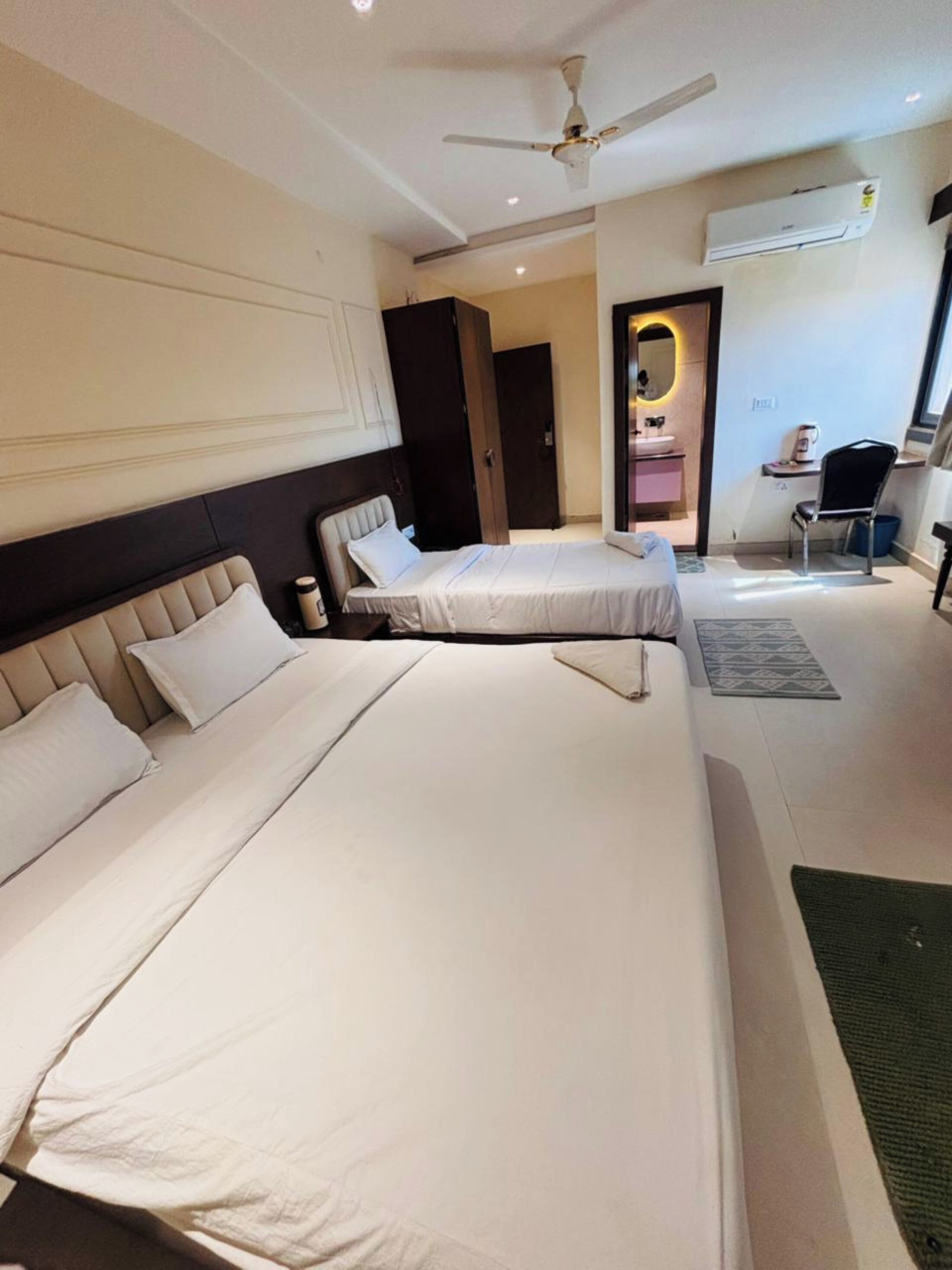 Habitación de hotel con dos camas, ventilador de techo, unidad de aire acondicionado, un pequeño escritorio con silla y un baño visible a través de una puerta abierta.